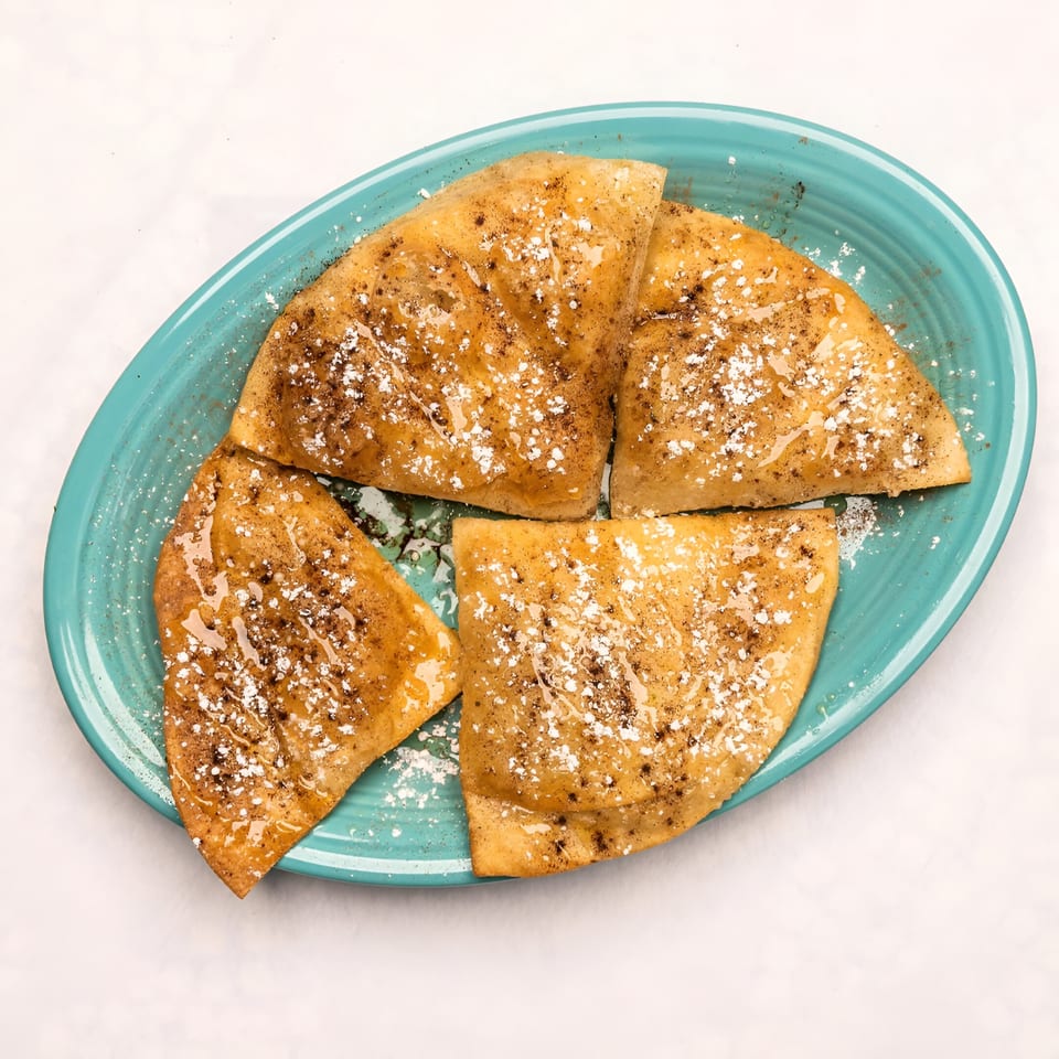 Four golden-brown sopapillas, drizzled with honey and sprinkled with powdered sugar, on a blue plate.