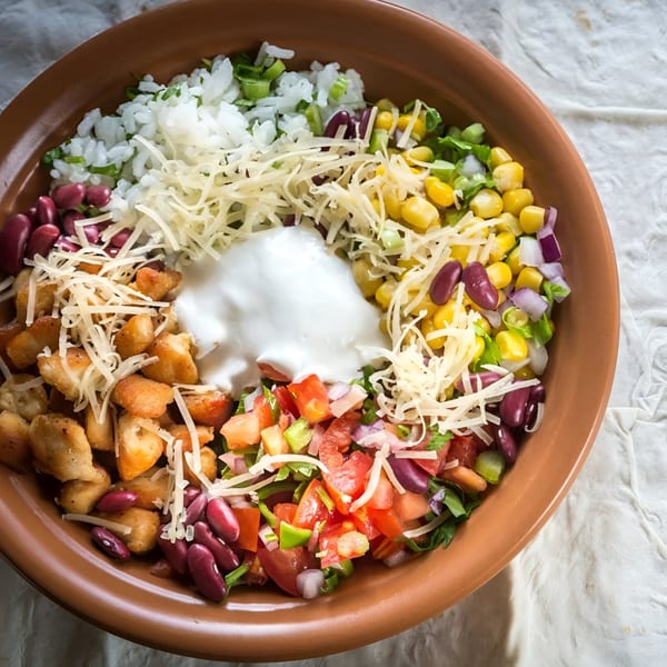 A colorful Mexican burrito bowl with chicken, rice, beans, corn, salsa, cheese, and sour cream.